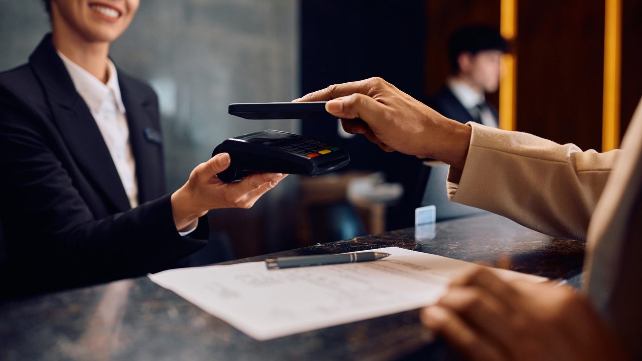 Hotel front-desk associate presenting a contactless payment terminal to a guest paying with a smartphone.